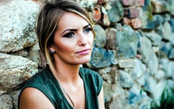 A woman with short blonde hair and wearing a green top sits against a stone wall, looking thoughtfully into the distance. The background features a mix of large and small stones.