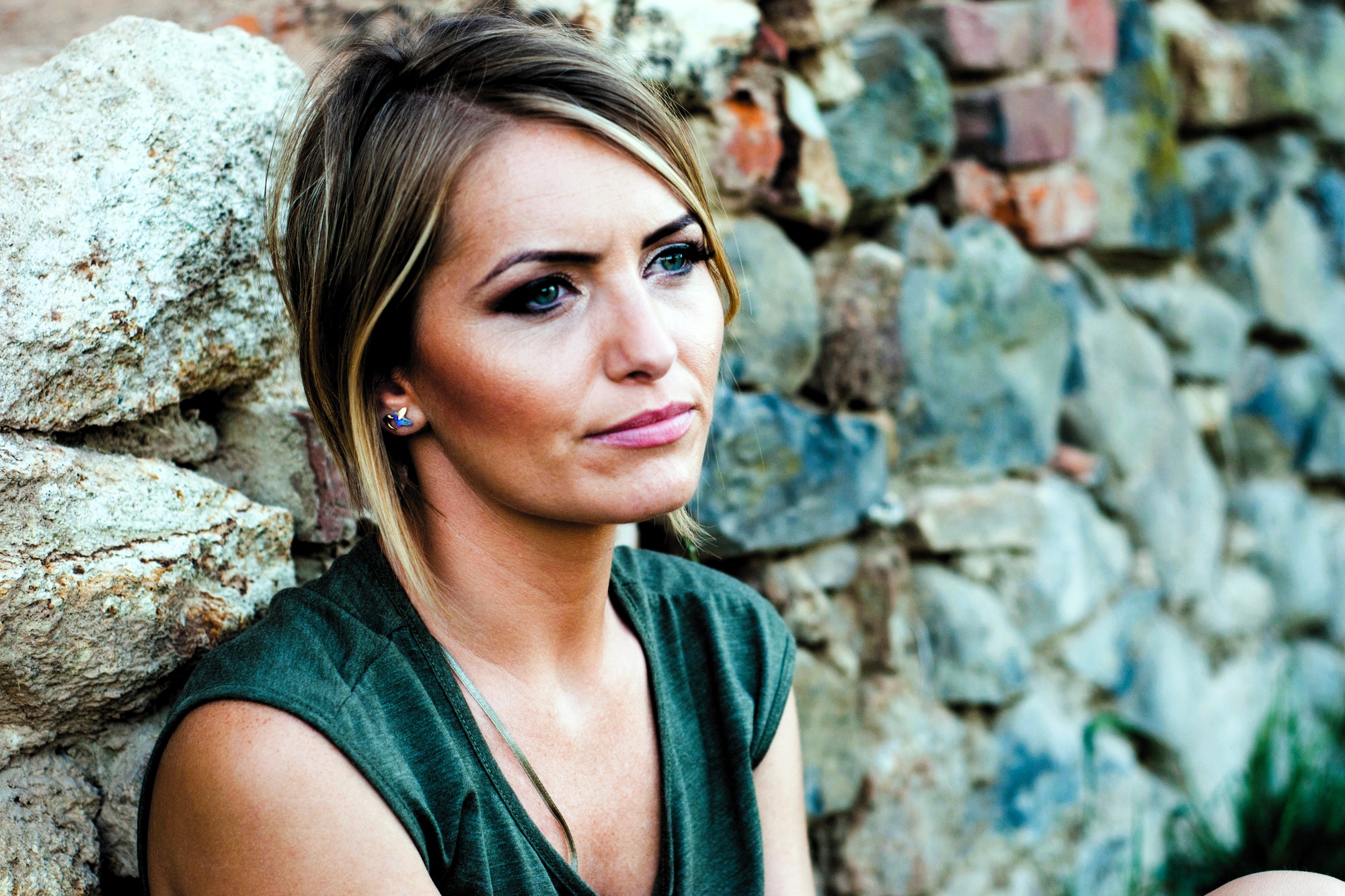 A woman with short blonde hair and wearing a green top sits against a stone wall, looking thoughtfully into the distance. The background features a mix of large and small stones.