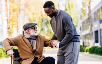 A young man helps an elderly man in a wheelchair. The elderly man is wearing a brown coat, scarf, cap, and sunglasses, while the young man is in a gray tracksuit. They are outdoors on a sunny day, surrounded by blurred greenery and buildings.