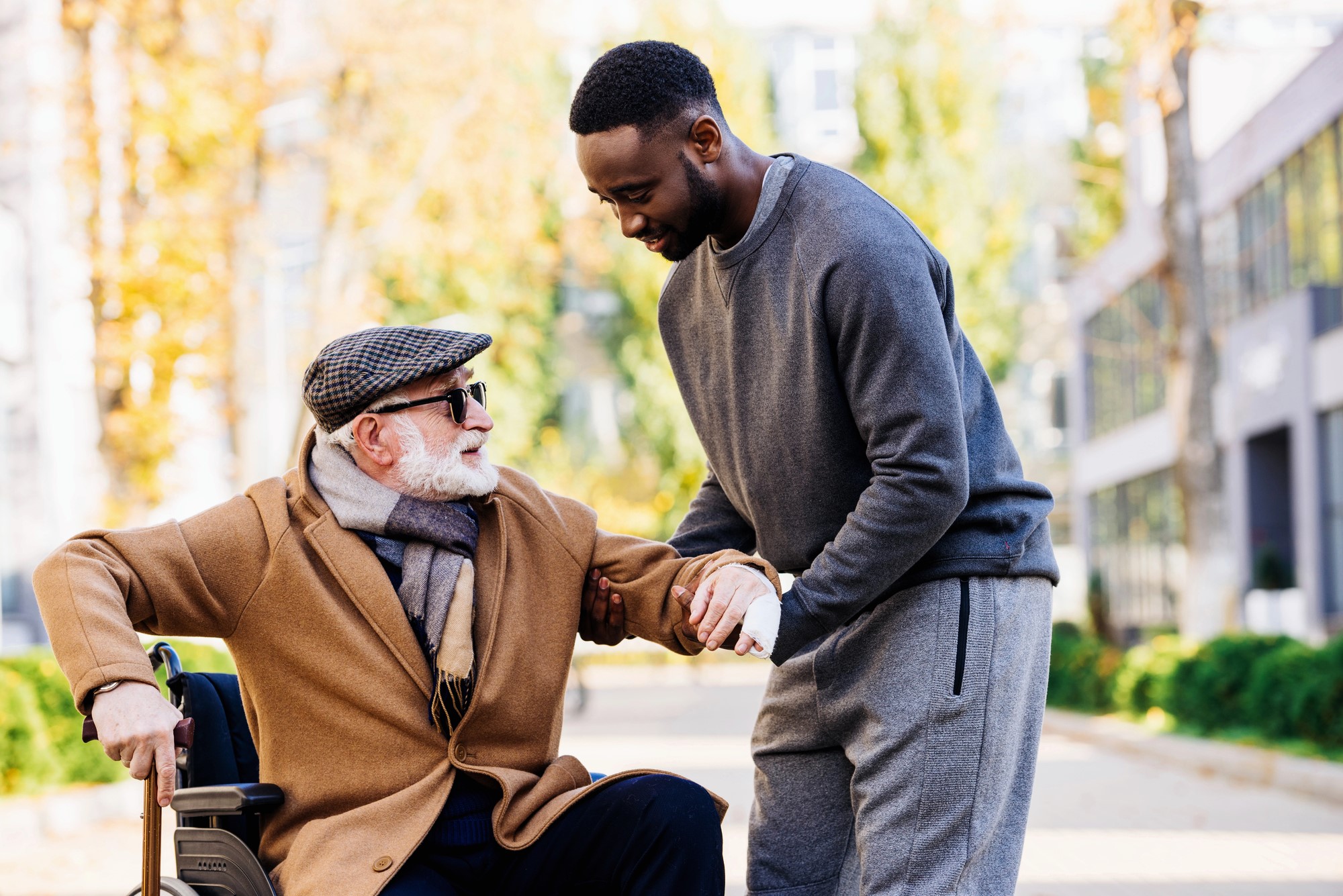 A young man helps an elderly man in a wheelchair. The elderly man is wearing a brown coat, scarf, cap, and sunglasses, while the young man is in a gray tracksuit. They are outdoors on a sunny day, surrounded by blurred greenery and buildings.