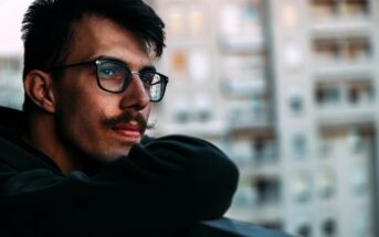 A person with glasses and a mustache leans on a railing, gazing thoughtfully to the side. They are wearing a dark hoodie. The background is a blurred view of apartment buildings.