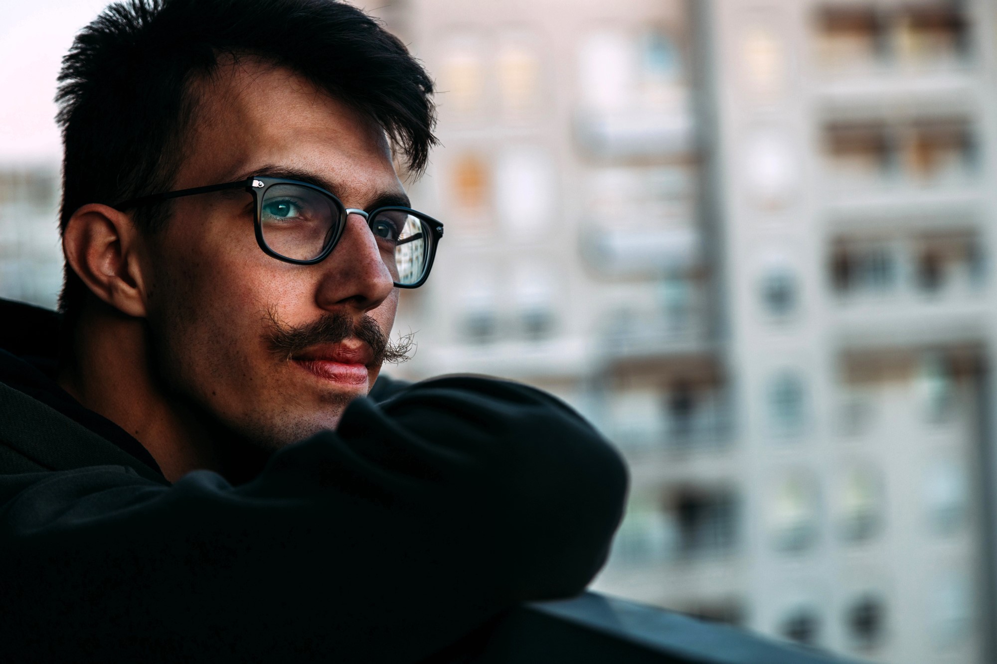 A person with glasses and a mustache leans on a railing, gazing thoughtfully to the side. They are wearing a dark hoodie. The background is a blurred view of apartment buildings.