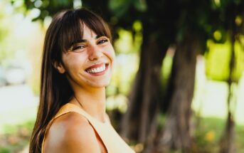 A woman with long brown hair and bangs smiles brightly while standing outdoors. She is wearing a sleeveless yellow top. The background features blurred greenery and trees, suggesting a sunny day in a park or garden.