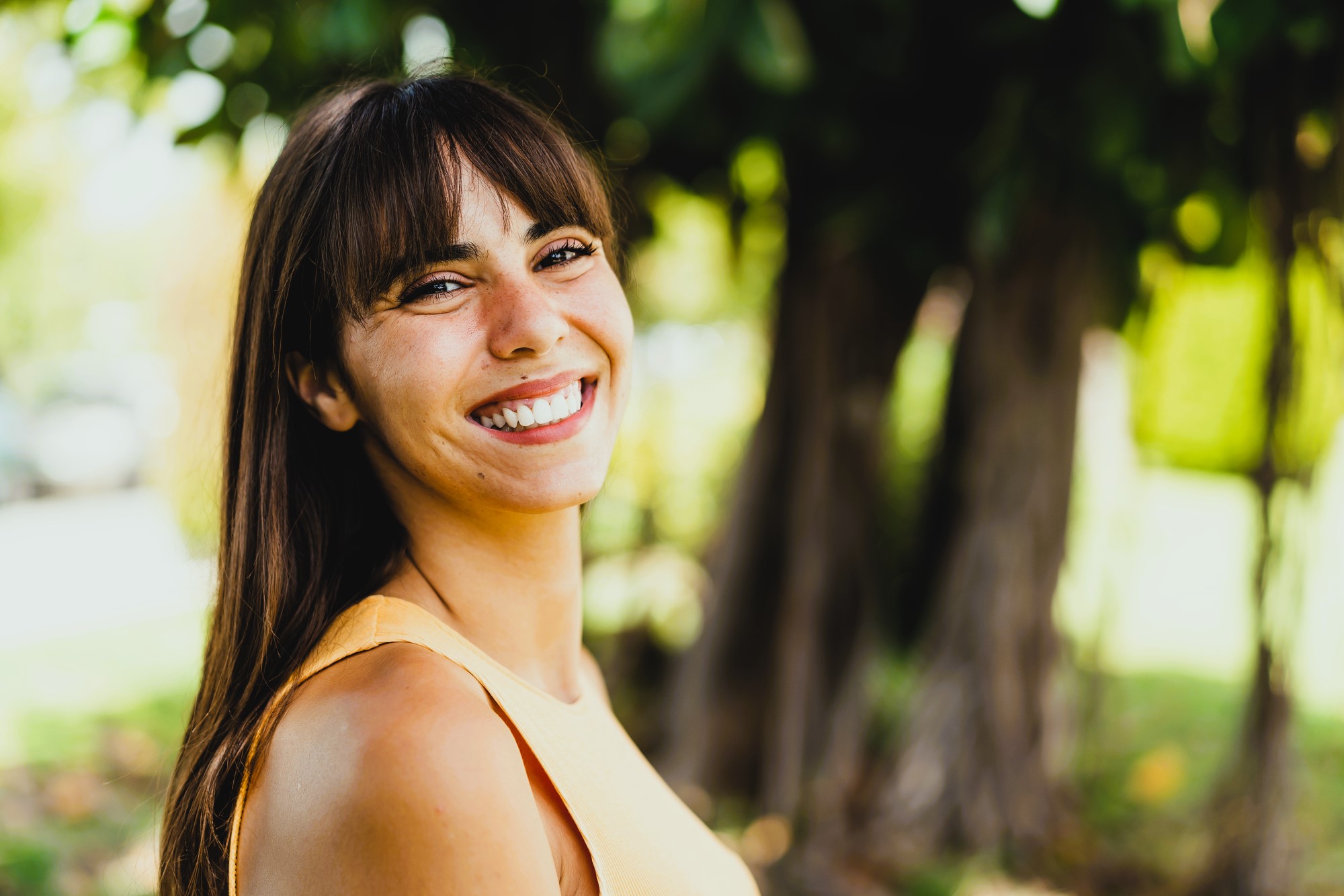 A woman with long brown hair and bangs smiles brightly while standing outdoors. She is wearing a sleeveless yellow top. The background features blurred greenery and trees, suggesting a sunny day in a park or garden.