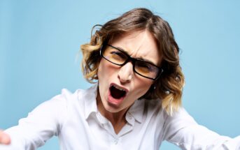 A woman with short, wavy hair and glasses is wearing a white shirt and appears to be yelling. She leans slightly forward against a blue background.