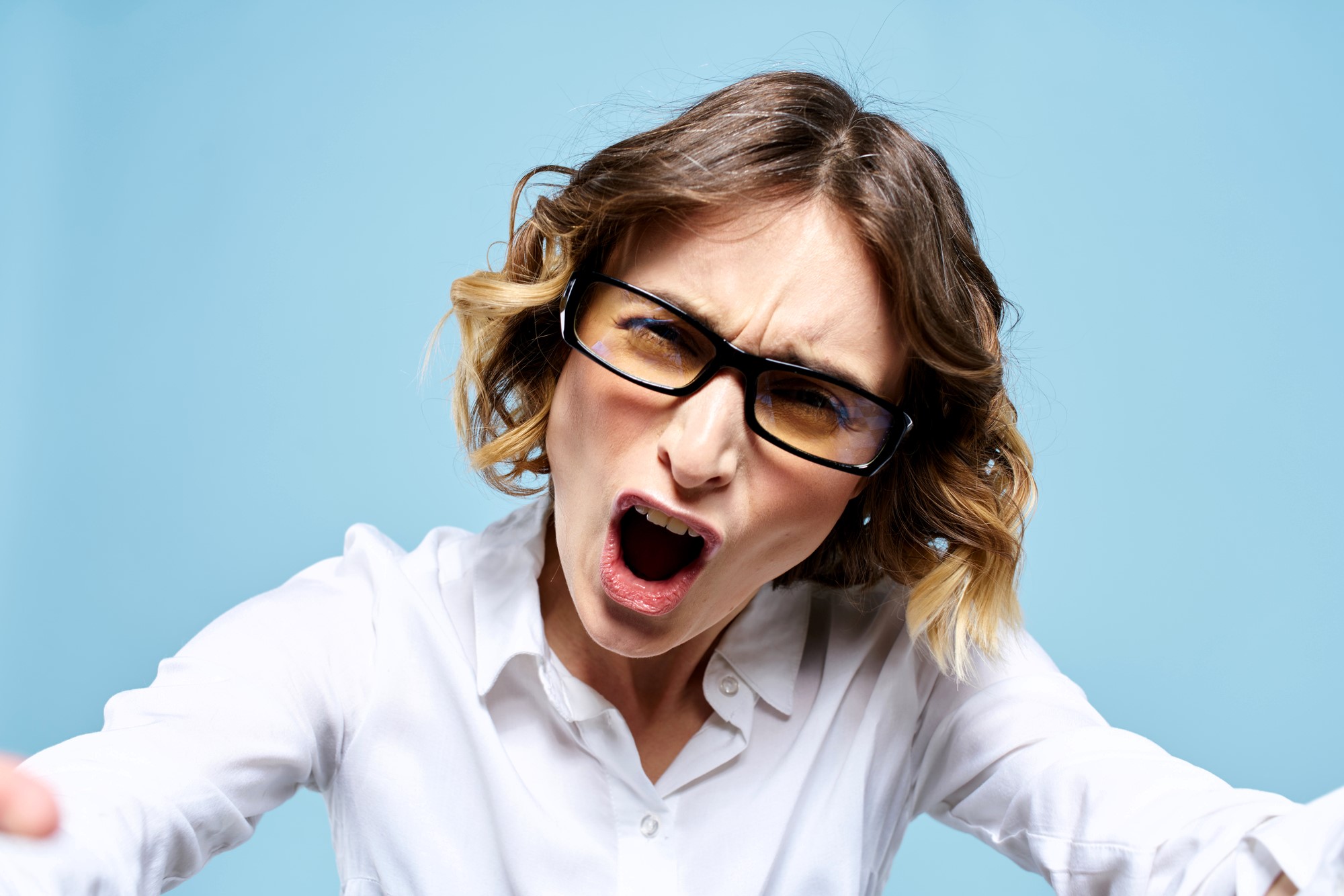 A woman with short, wavy hair and glasses is wearing a white shirt and appears to be yelling. She leans slightly forward against a blue background.