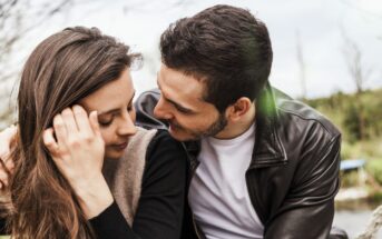 A couple sitting close together outdoors. The man is wearing a leather jacket and white shirt, looking lovingly at the woman, who is smiling with her head slightly turned. She has long brown hair and a relaxed expression. Trees and grass are in the background.