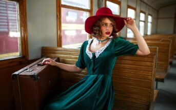 A woman in a vintage green dress and red hat sits on a wooden train bench, with a vintage suitcase beside her. She gazes out the window, while the train's wooden interior and an outside train car are visible.