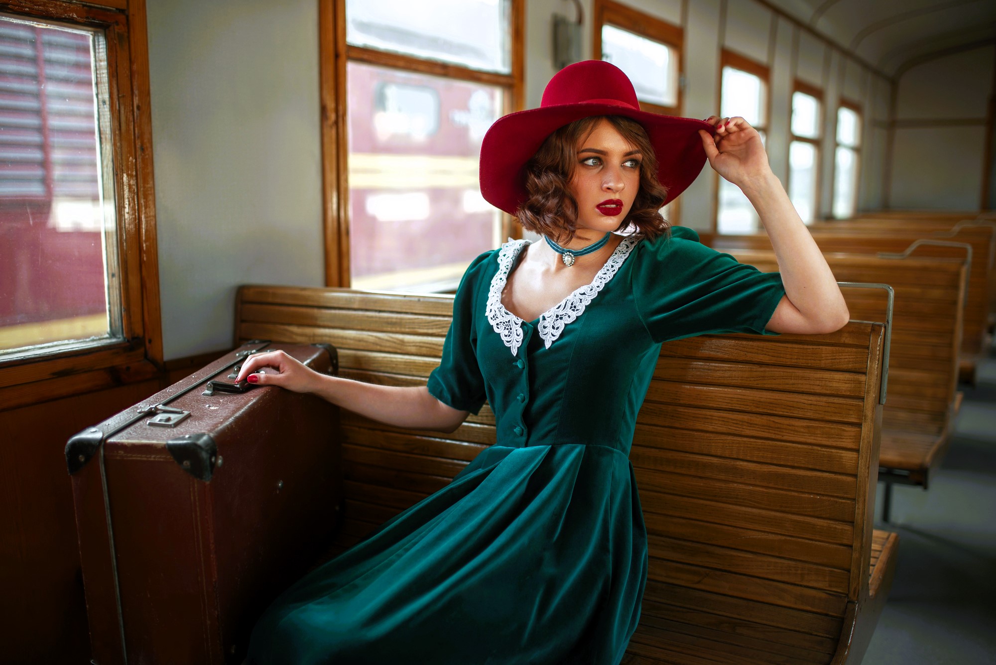 A woman in a vintage green dress and red hat sits on a wooden train bench, with a vintage suitcase beside her. She gazes out the window, while the train's wooden interior and an outside train car are visible.