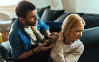 A man and woman are sitting on a sofa, engaged in a tense conversation. The man gestures with open hands, appearing to explain or plead, while the woman turns away with a closed expression, avoiding eye contact.