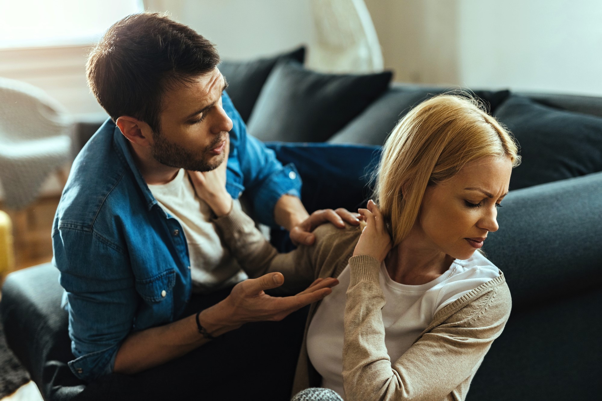 A man and woman are sitting on a sofa, engaged in a tense conversation. The man gestures with open hands, appearing to explain or plead, while the woman turns away with a closed expression, avoiding eye contact.