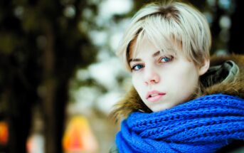 Person with short blonde hair wearing a blue scarf and a fur-trimmed coat, standing outdoors. The background is blurred, possibly depicting trees or a park setting, creating a cold, wintry atmosphere.