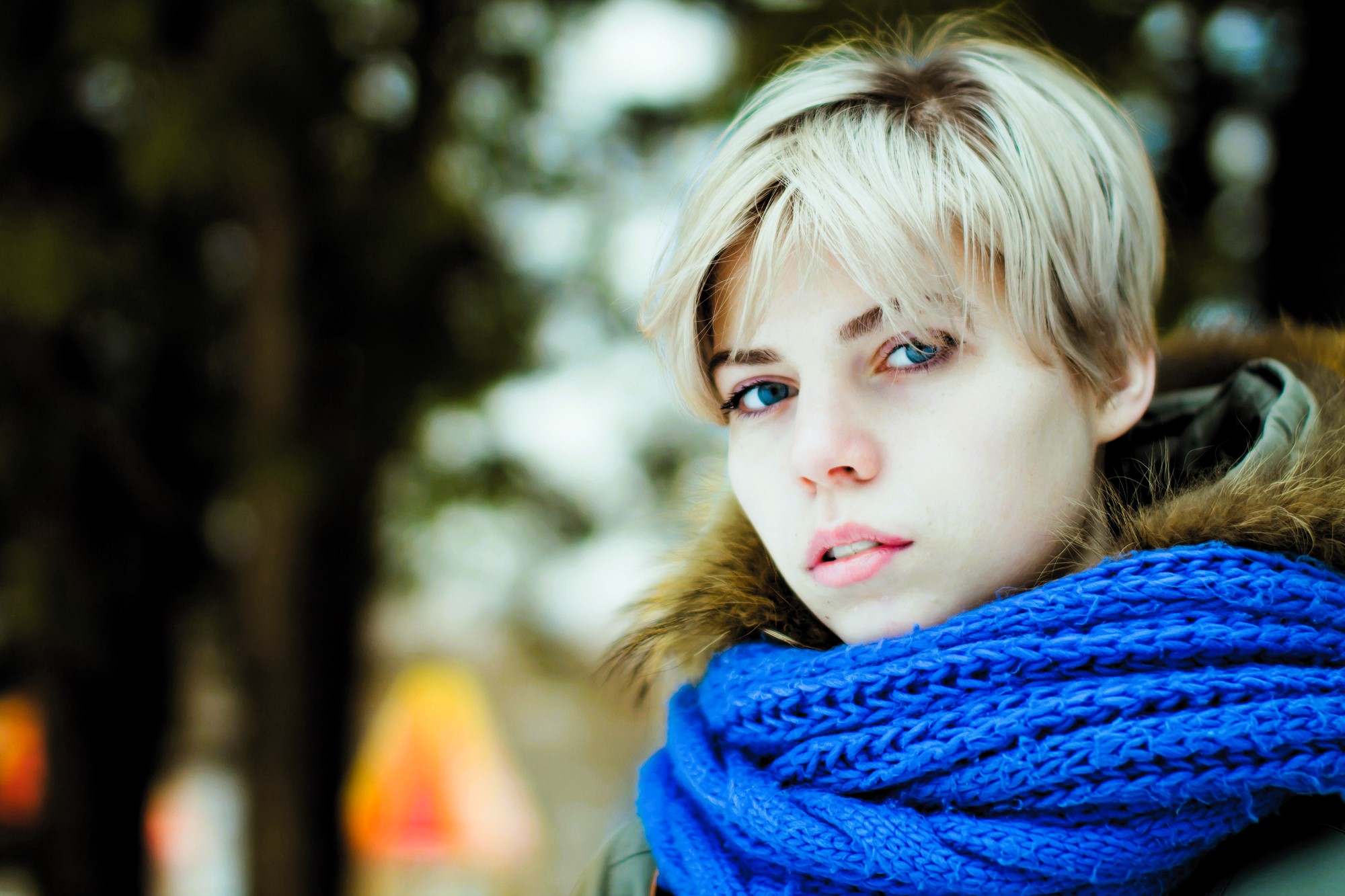 Person with short blonde hair wearing a blue scarf and a fur-trimmed coat, standing outdoors. The background is blurred, possibly depicting trees or a park setting, creating a cold, wintry atmosphere.