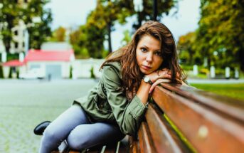 Woman with long hair and a green jacket, leaning on a wooden bench in an outdoor setting. She is looking directly at the camera, with trees, buildings, and a cloudy sky in the background.