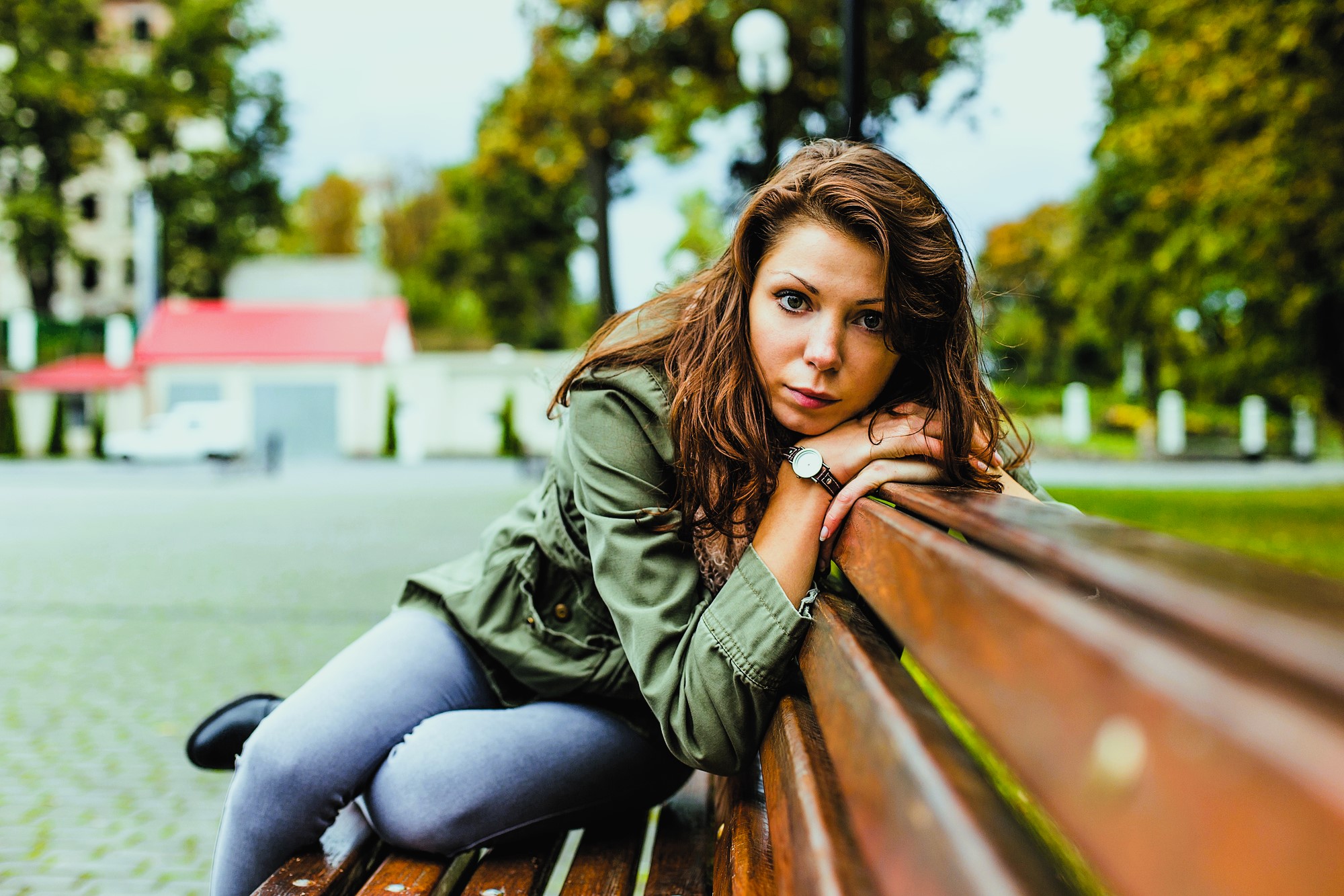 Woman with long hair and a green jacket, leaning on a wooden bench in an outdoor setting. She is looking directly at the camera, with trees, buildings, and a cloudy sky in the background.