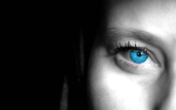 Close-up of an eye with a striking blue iris, surrounded by grayscale skin. The image is partially shadowed, emphasizing the eye's vivid color and detail. The lighting highlights the texture of the skin and eyelashes.