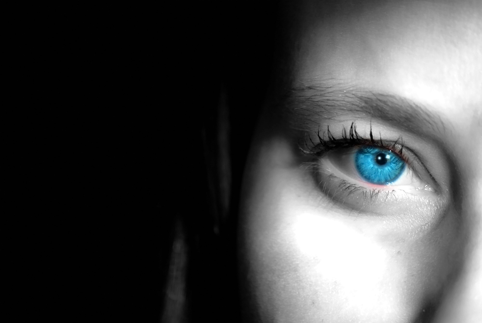 Close-up of an eye with a striking blue iris, surrounded by grayscale skin. The image is partially shadowed, emphasizing the eye's vivid color and detail. The lighting highlights the texture of the skin and eyelashes.