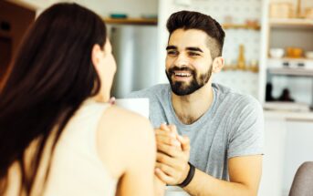 A man with a beard and dark hair smiles warmly at a woman with long dark hair. They are sitting indoors, holding hands. The background shows a kitchen with shelves and various kitchen items.