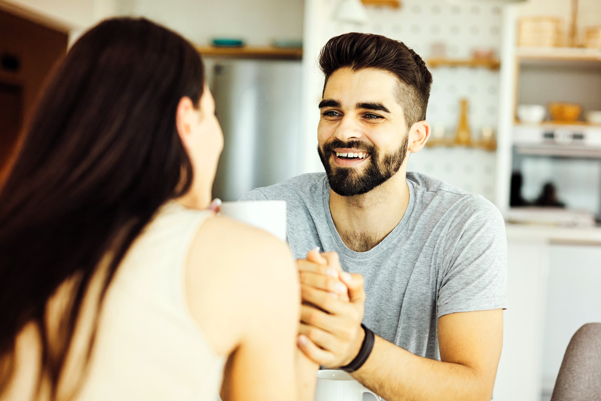 A man with a beard and dark hair smiles warmly at a woman with long dark hair. They are sitting indoors, holding hands. The background shows a kitchen with shelves and various kitchen items.