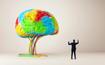 A man in a suit stands with raised hands, facing a large, colorful brain sculpture. The brain, placed against a beige background, features vibrant swirls of blue, green, red, and yellow, with wires emerging from its base.