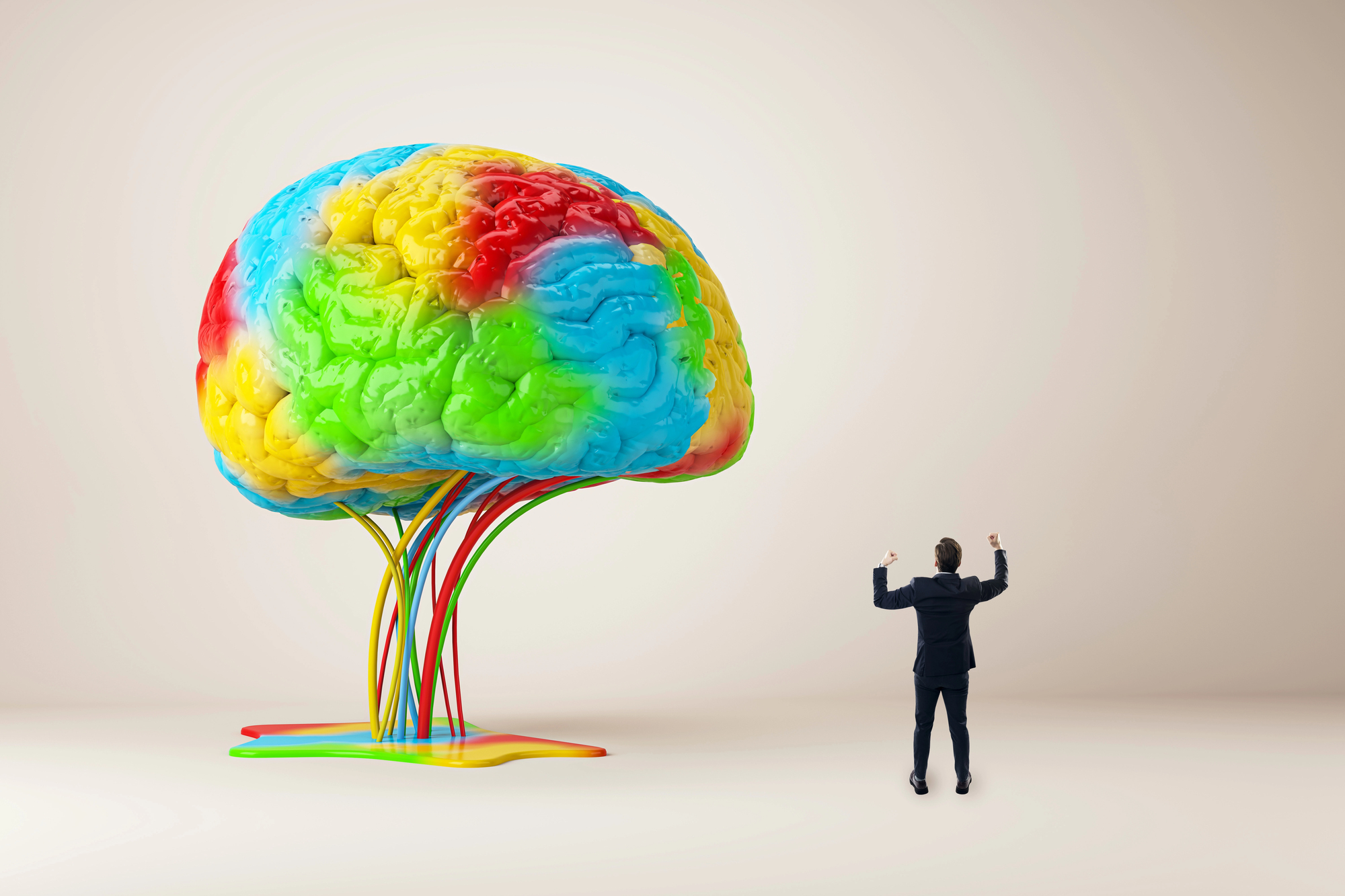 A man in a suit stands with raised hands, facing a large, colorful brain sculpture. The brain, placed against a beige background, features vibrant swirls of blue, green, red, and yellow, with wires emerging from its base.