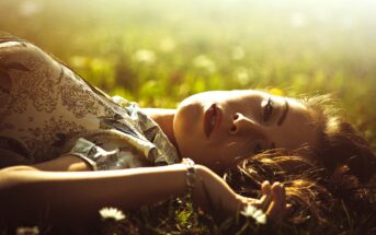 A woman lies on grass, gazing upward with a serene expression. Sunlight bathes the scene, casting a warm glow. She wears a floral-patterned top and a bracelet, surrounded by blurred white flowers in a tranquil setting.