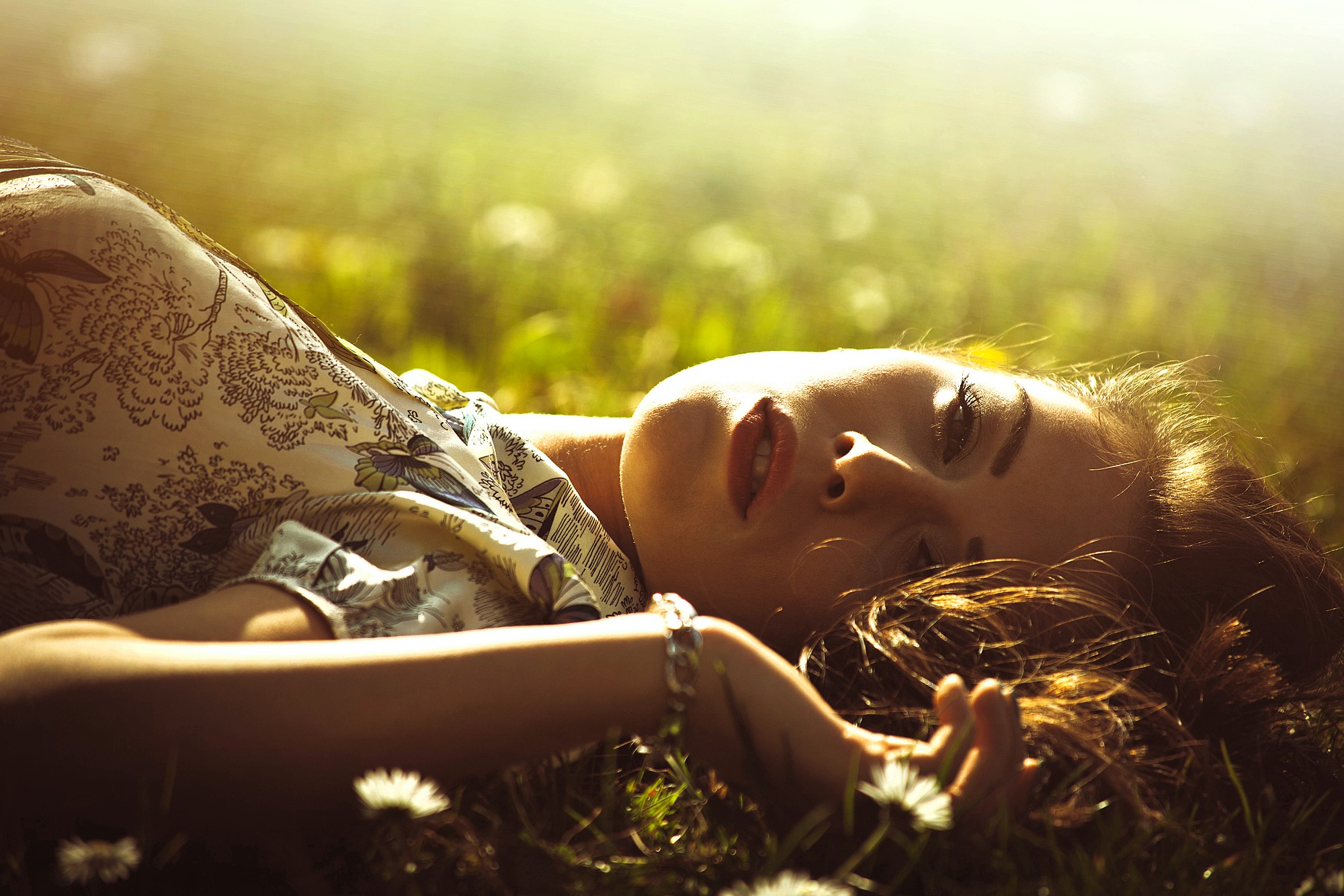 A woman lies on grass, gazing upward with a serene expression. Sunlight bathes the scene, casting a warm glow. She wears a floral-patterned top and a bracelet, surrounded by blurred white flowers in a tranquil setting.