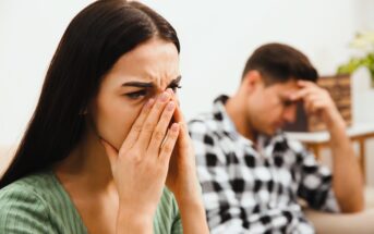 A woman with long dark hair covers her face with her hands, appearing upset. A man in a checkered shirt sits in the background, resting his head on his hand, looking troubled. The setting is a softly lit room.