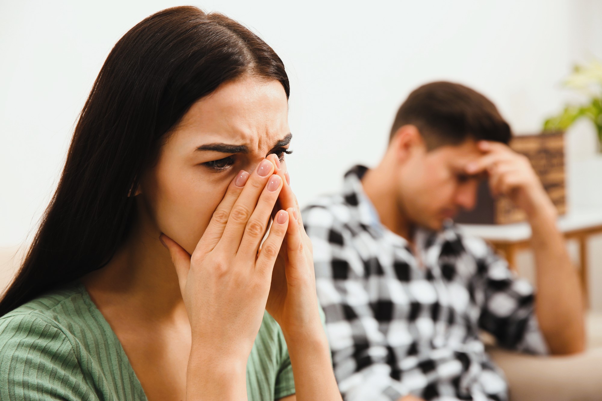 A woman with long dark hair covers her face with her hands, appearing upset. A man in a checkered shirt sits in the background, resting his head on his hand, looking troubled. The setting is a softly lit room.