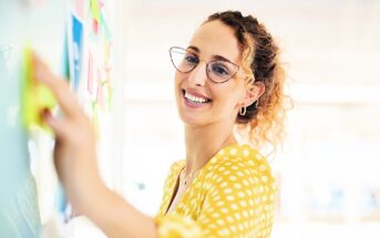 A woman in a yellow polka dot blouse smiles while placing sticky notes on a board. She wears glasses and has curly hair, which is tied back. The background is softly blurred, suggesting a bright, open environment.