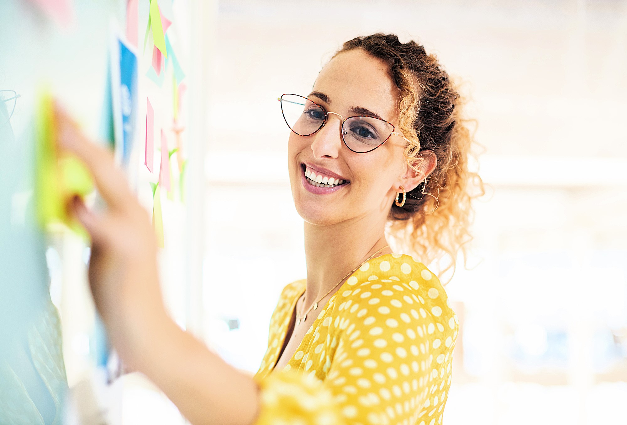 A woman in a yellow polka dot blouse smiles while placing sticky notes on a board. She wears glasses and has curly hair, which is tied back. The background is softly blurred, suggesting a bright, open environment.