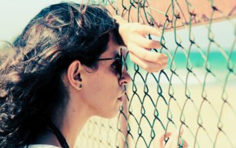 A woman with long, wavy hair and sunglasses leans against a chain-link fence, gazing out at a blurry beach and ocean. Her hand rests on the fence, and she wears a white shirt and stud earrings. The scene is bright and sunlit.