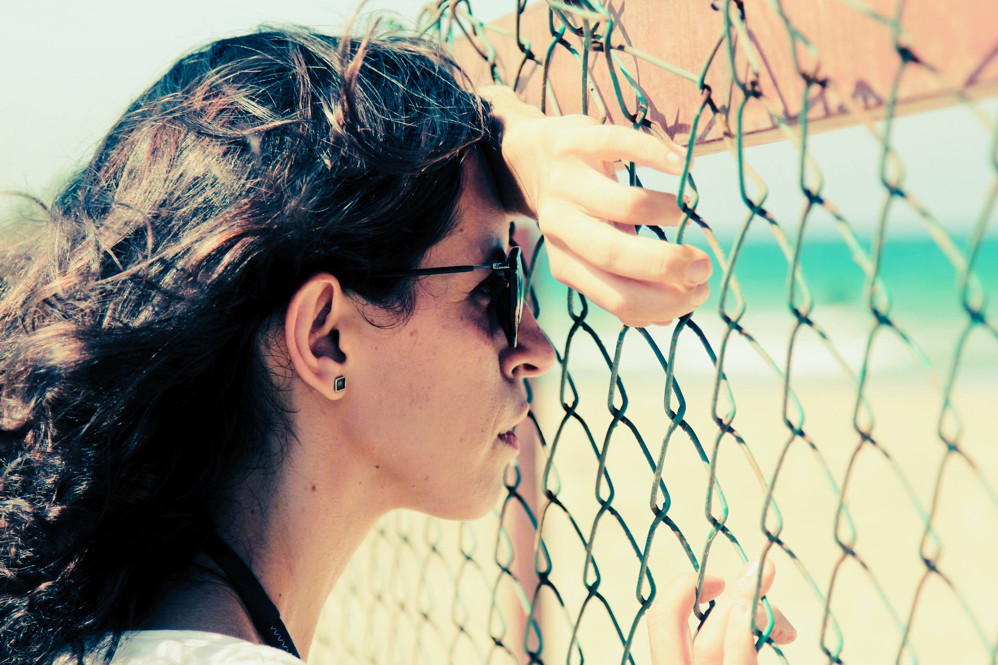 A woman with long, wavy hair and sunglasses leans against a chain-link fence, gazing out at a blurry beach and ocean. Her hand rests on the fence, and she wears a white shirt and stud earrings. The scene is bright and sunlit.