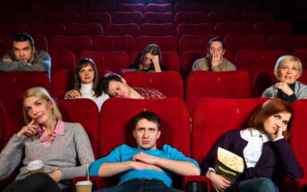 A group of people sitting in a theater with red seats, looking bored or sleepy. Some hold snacks and drinks, while others rest their heads on their hands or lean back. The theater is mostly empty, and their expressions suggest disinterest.