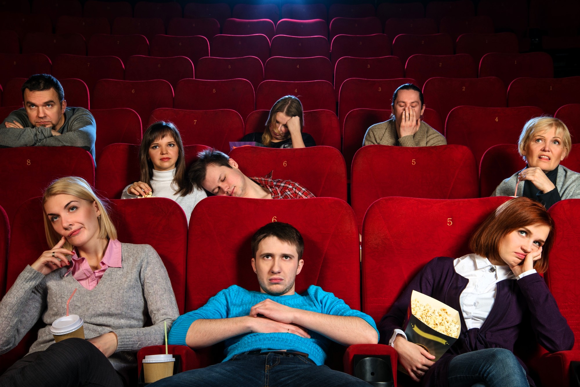 A group of people sitting in a theater with red seats, looking bored or sleepy. Some hold snacks and drinks, while others rest their heads on their hands or lean back. The theater is mostly empty, and their expressions suggest disinterest.