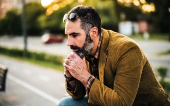 A man with a beard sits outdoors on a bench, leaning forward with hands clasped near his face. He wears a brown jacket, checked shirt, jeans, and sunglasses resting on his head. The background is a blurred street scene with trees.