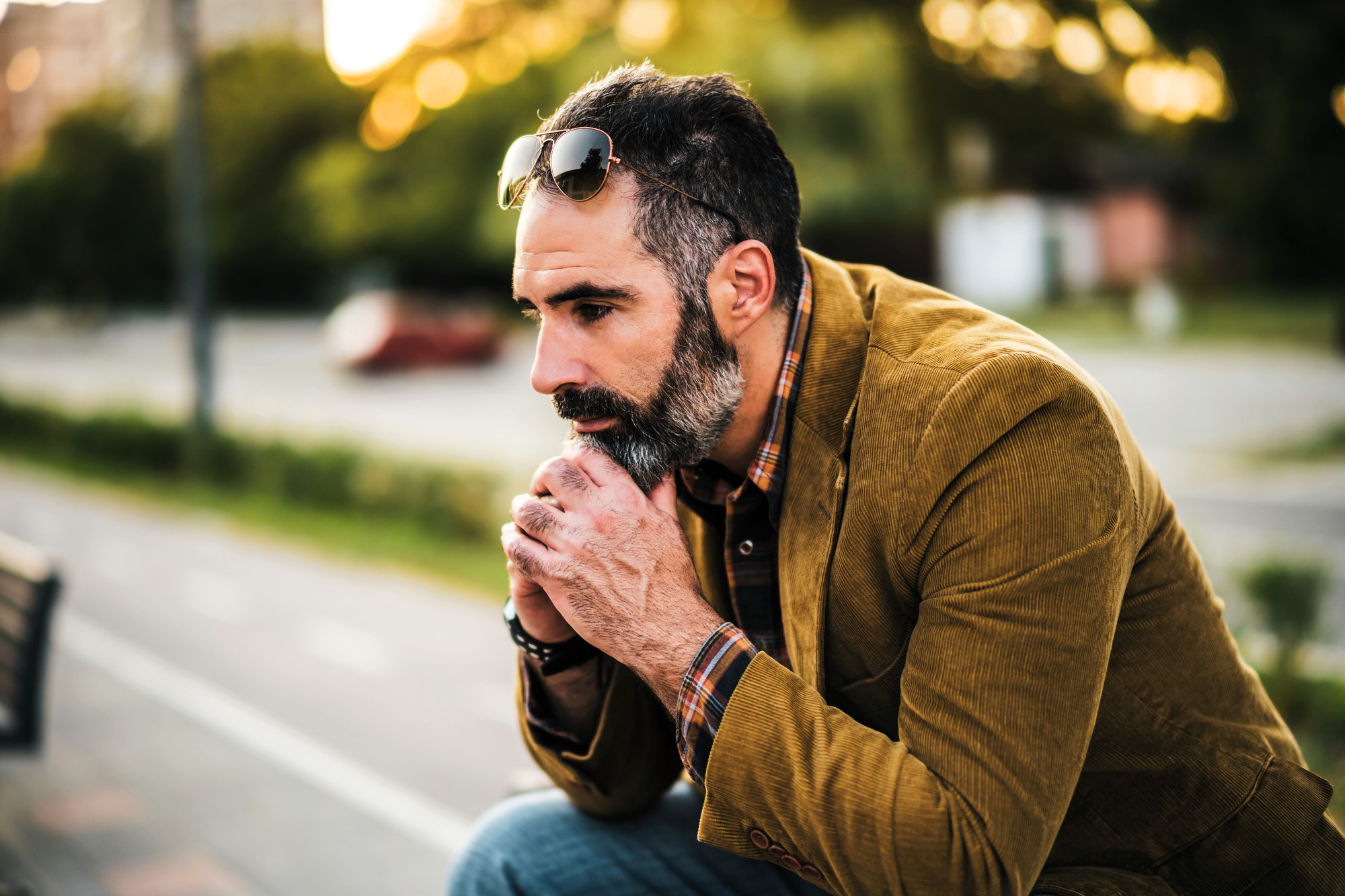 A man with a beard sits outdoors on a bench, leaning forward with hands clasped near his face. He wears a brown jacket, checked shirt, jeans, and sunglasses resting on his head. The background is a blurred street scene with trees.