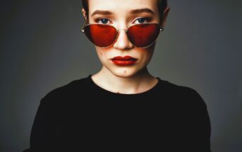 Person with short hair wearing red-tinted sunglasses and red lipstick, dressed in a black top. The background is a plain, dark gray, and they are looking directly at the camera with a neutral expression.