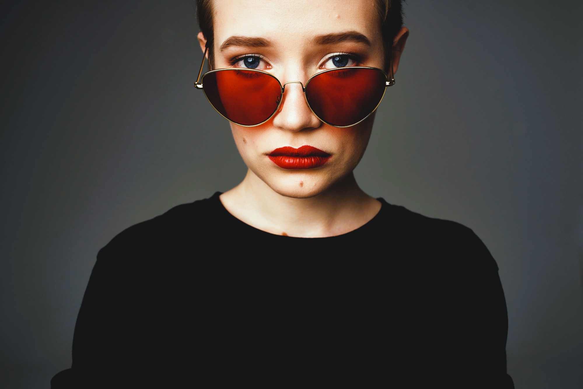 Person with short hair wearing red-tinted sunglasses and red lipstick, dressed in a black top. The background is a plain, dark gray, and they are looking directly at the camera with a neutral expression.