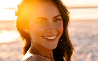 A woman with long brown hair and a warm smile stands on a beach at sunset. Sunlight creates a golden glow around her, highlighting her natural features against the blurred sandy background.