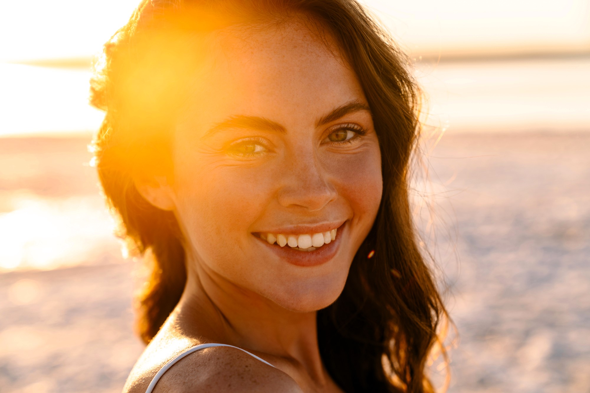 A woman with long brown hair and a warm smile stands on a beach at sunset. Sunlight creates a golden glow around her, highlighting her natural features against the blurred sandy background.