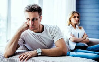 A man in a white t-shirt sits on a couch with his head resting on his hand, looking away with a thoughtful expression. A woman sits in the background, arms crossed, appearing distant. The room is well-lit with soft natural light.