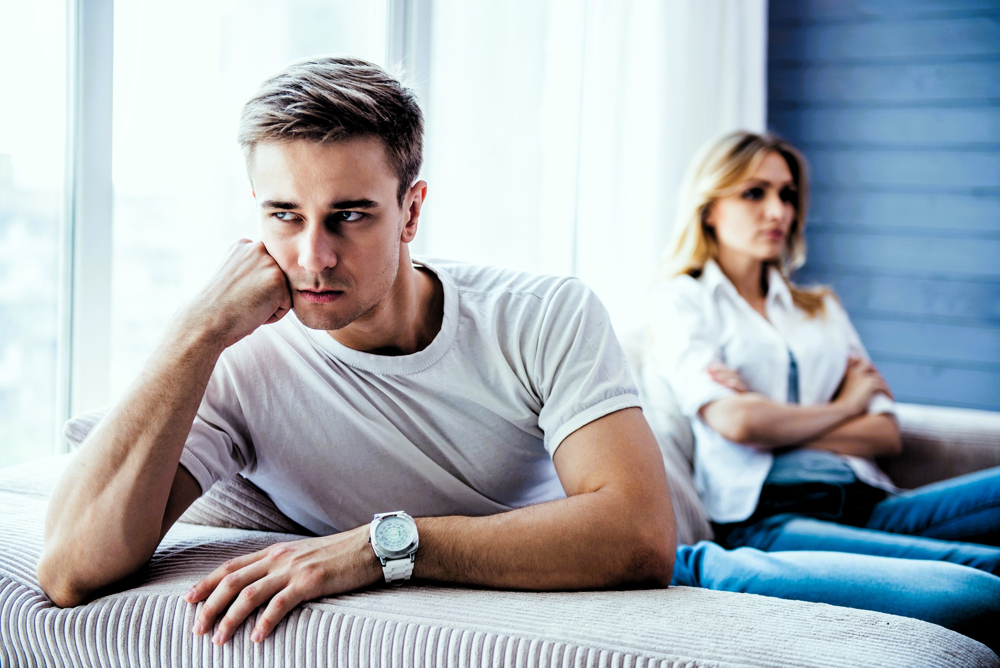 A man in a white t-shirt sits on a couch with his head resting on his hand, looking away with a thoughtful expression. A woman sits in the background, arms crossed, appearing distant. The room is well-lit with soft natural light.