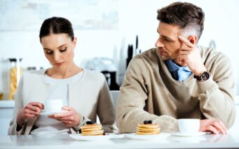 A man and woman sit at a kitchen counter with stacks of pancakes and cups. The woman looks at her cup while holding it, and the man looks at her with his hand on his face, slightly frowning. The setting is bright and modern.