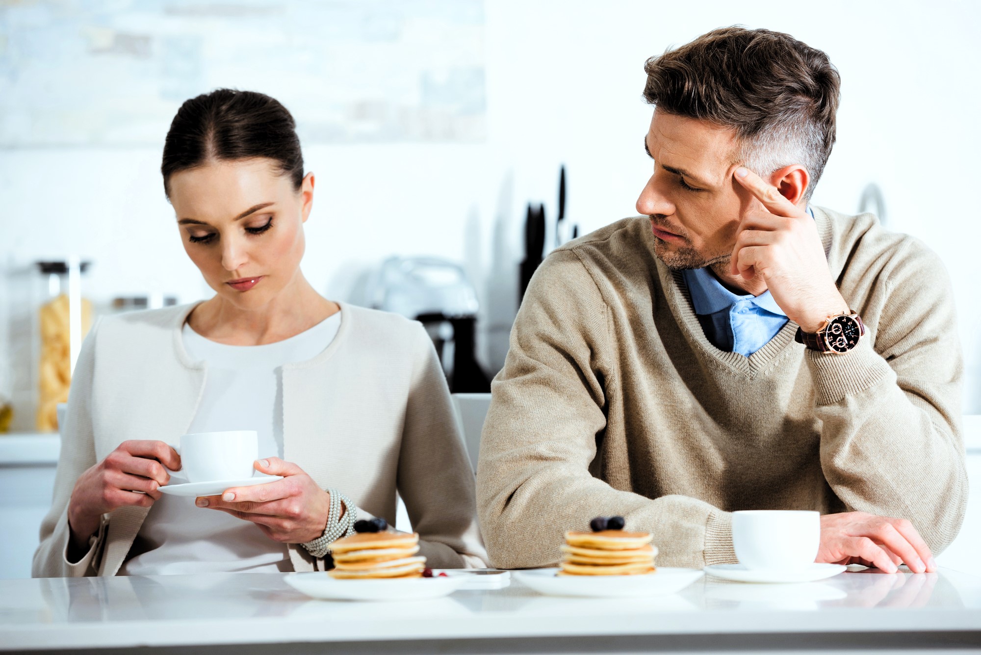 A man and woman sit at a kitchen counter with stacks of pancakes and cups. The woman looks at her cup while holding it, and the man looks at her with his hand on his face, slightly frowning. The setting is bright and modern.