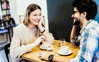 A woman and a man sit at a wooden table in a café, smiling and chatting. They both have coffee in front of them. She wears a light sweater, and he wears a plaid shirt and glasses. Glasses of water are also on the table.