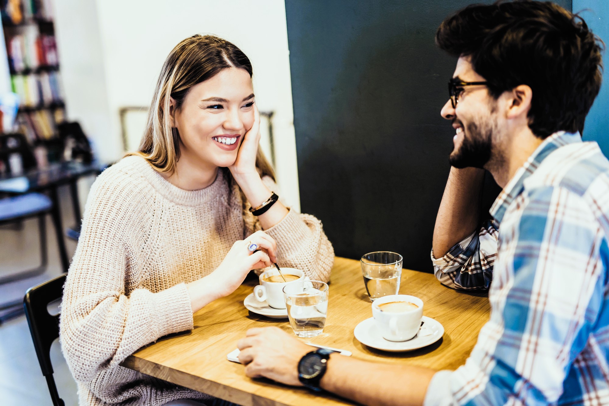 A woman and a man sit at a wooden table in a café, smiling and chatting. They both have coffee in front of them. She wears a light sweater, and he wears a plaid shirt and glasses. Glasses of water are also on the table.