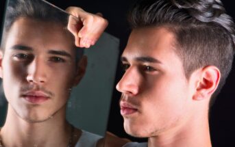 A young man with dark hair looks into a mirror, his reflection showing a pensive expression. He holds the mirror up with his left hand, which is slightly visible. The background is dark, emphasizing the contrast between the man and his reflection.