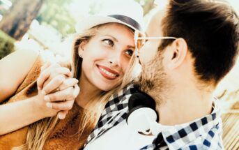 A smiling couple sit closely, gazing at each other. The woman wears a straw hat and holds the man's hand, while the man, with glasses, has headphones around his neck. They are outdoors with sunlight filtering through trees.