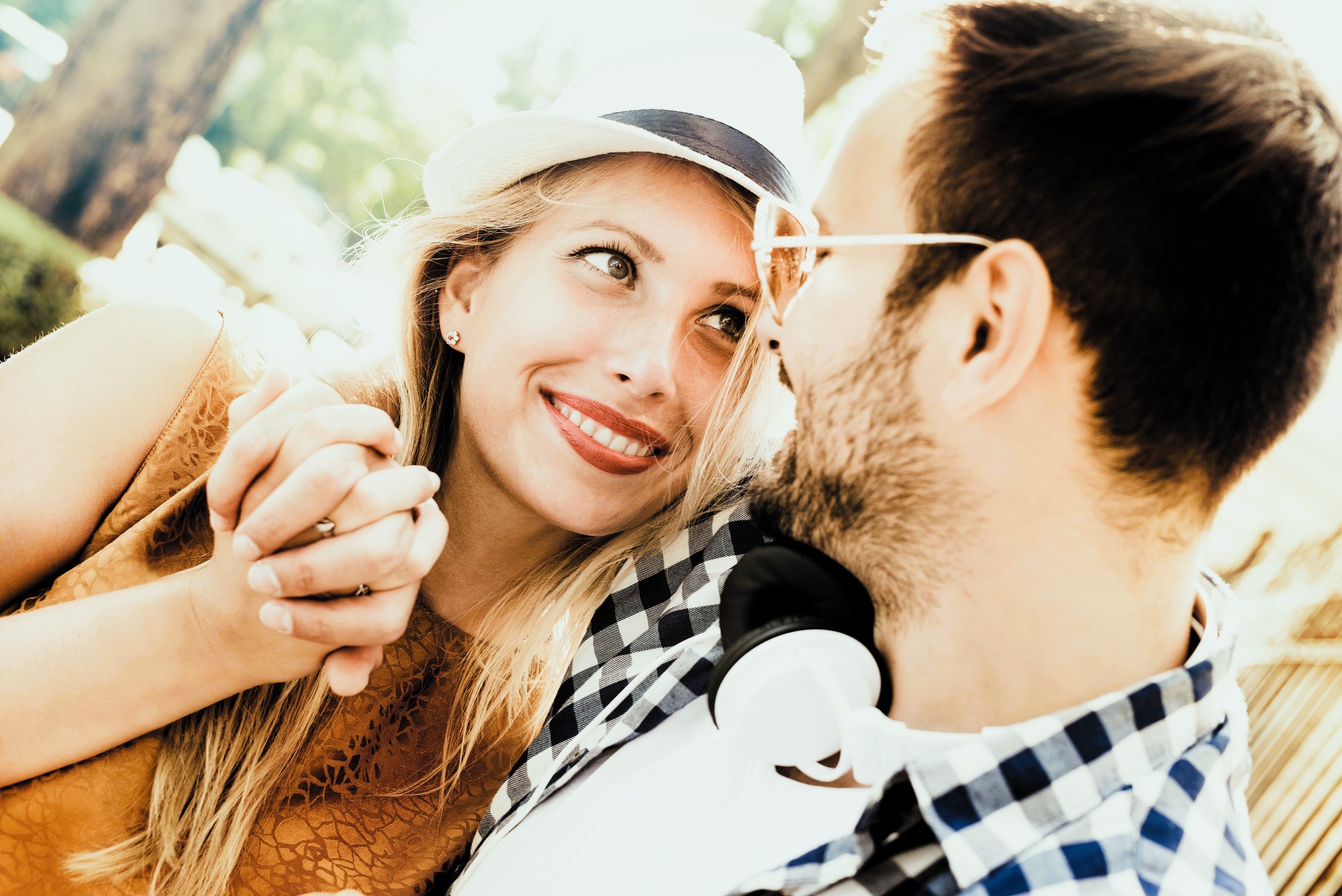 A smiling couple sit closely, gazing at each other. The woman wears a straw hat and holds the man's hand, while the man, with glasses, has headphones around his neck. They are outdoors with sunlight filtering through trees.